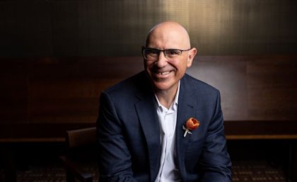 UQ 2023 Alumnus of the Year Professor George Mellick. A man in a navy suit with a red flower in his buttonhole smiles broadly.
